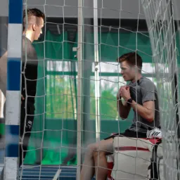 Dwóch młodych mężczyzn w hali sportowej, jeden stoi oparty o słupek bramki, drugi siedzi na ławce za siatką bramki, zakładając bandaże na nadgarstki.