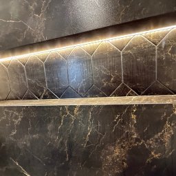 Płytkarz Śrem - Ciemna, marmurkowa ściana z podświetlaną wnęką wyłożoną heksagonalnymi płytkami. Widoczne złote żyłki w marmurze i precyzyjne fugi.