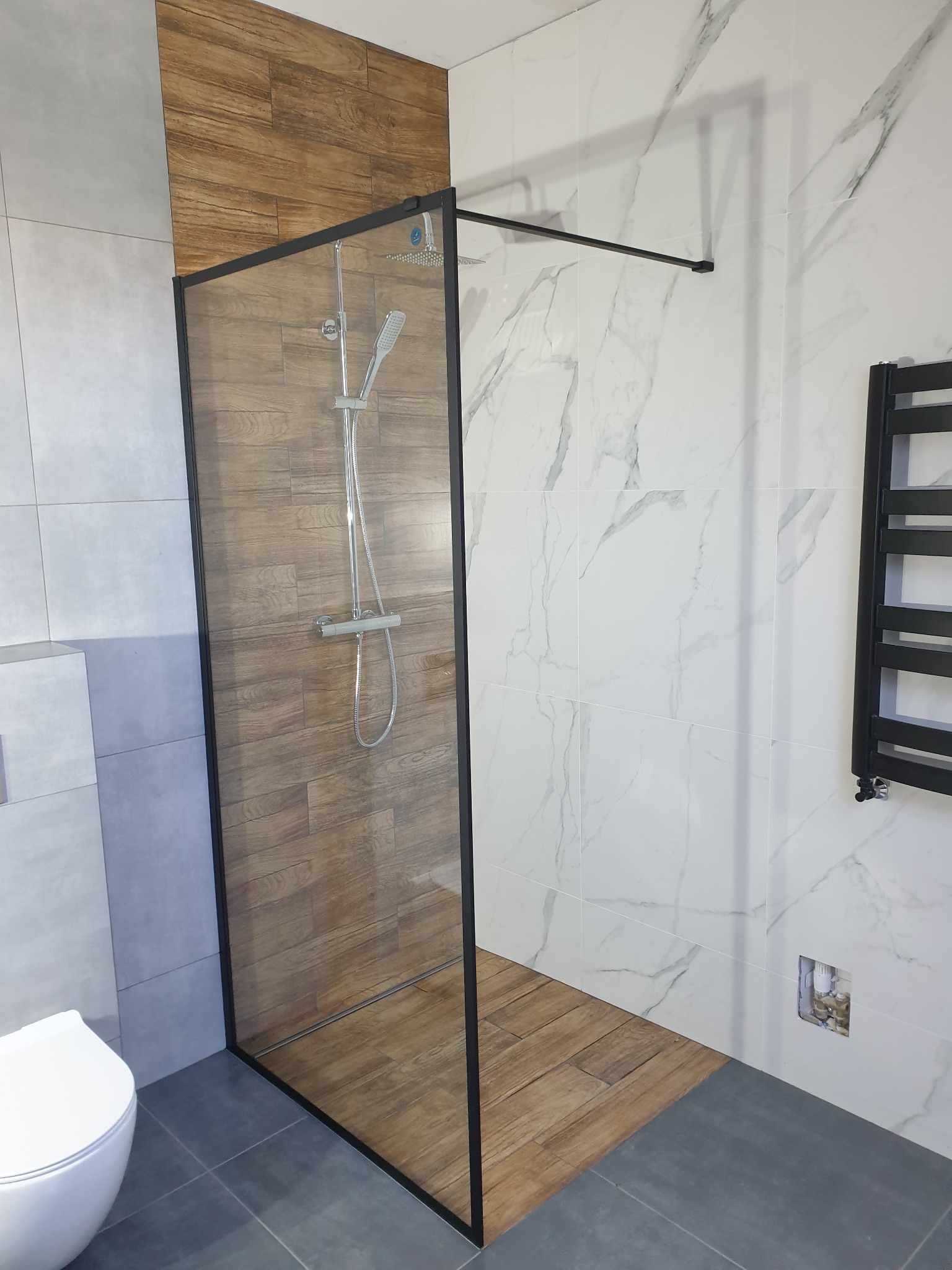 Nowoczesna kabina prysznicowa walk-in z czarną ramą, drewnopodobne płytki wewnątrz kabiny, marmurowe płytki na ścianach, szary gres na podłodze, widoczny grzejnik drabinkowy i toaleta.