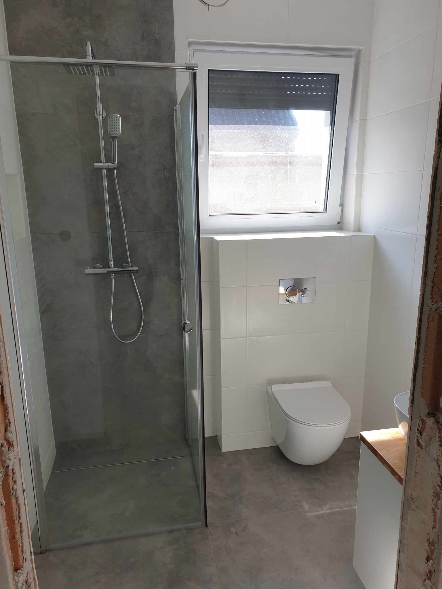 Nowoczesna łazienka w trakcie wykończenia, z widoczną kabiną prysznicową z szarą ścianą, podwieszaną toaletą, białymi płytkami i fragmentem niezabezpieczonej ściany w surowym stanie.