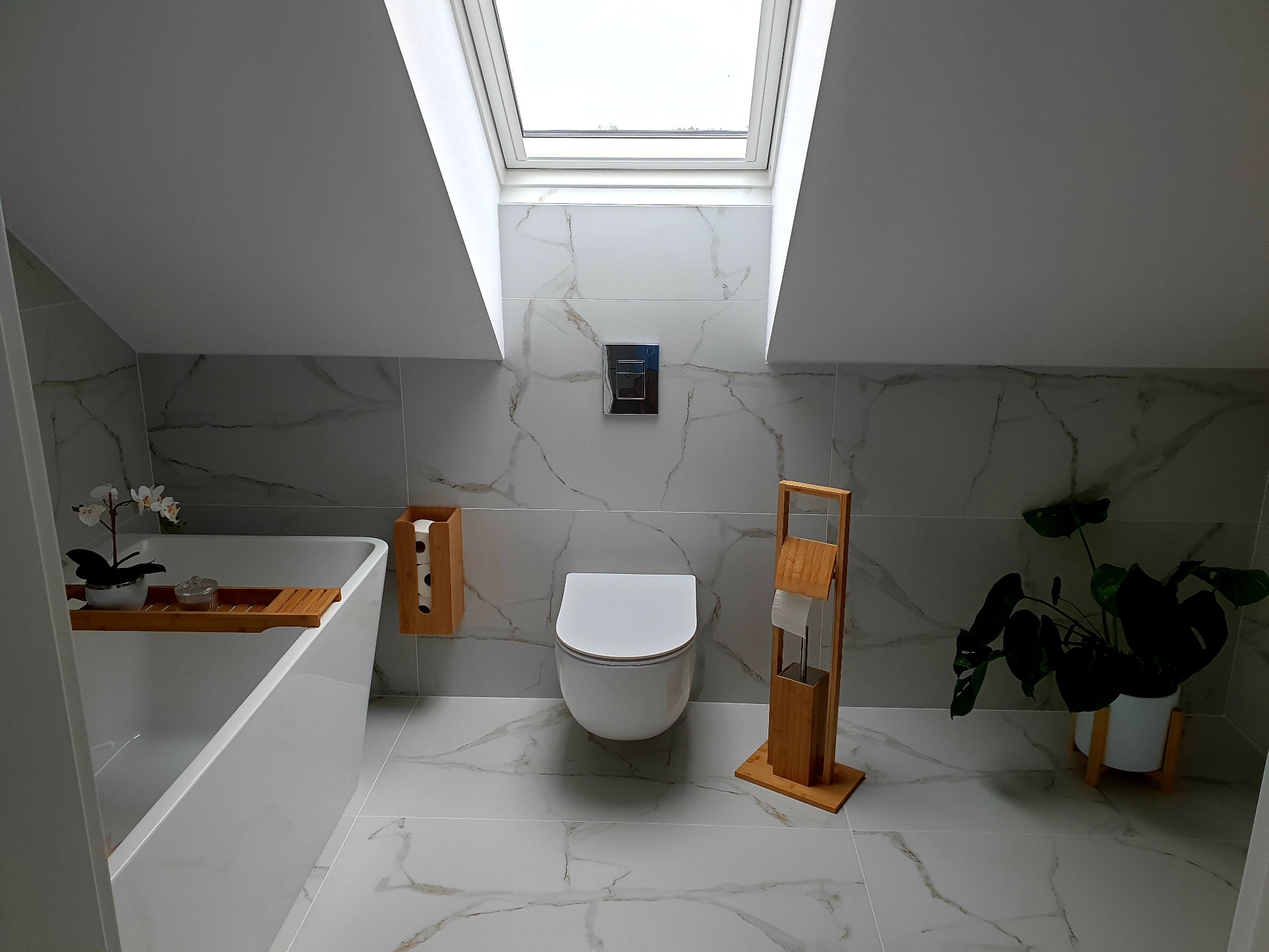 Nowoczesna łazienka na poddaszu z białymi płytkami imitującymi marmur, podwieszaną toaletą, drewnianymi akcesoriami łazienkowymi i roślinami doniczkowymi, oświetlona światłem wpadającym przez okno...