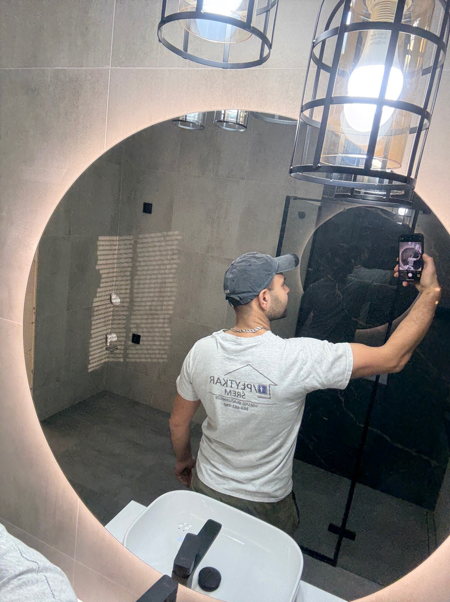 Okrągłe lustro z podświetleniem LED odbija wykończoną łazienkę z szarymi płytkami i czarnymi akcentami. Mężczyzna robi zdjęcie telefonem, widoczna umywalka i lampa.