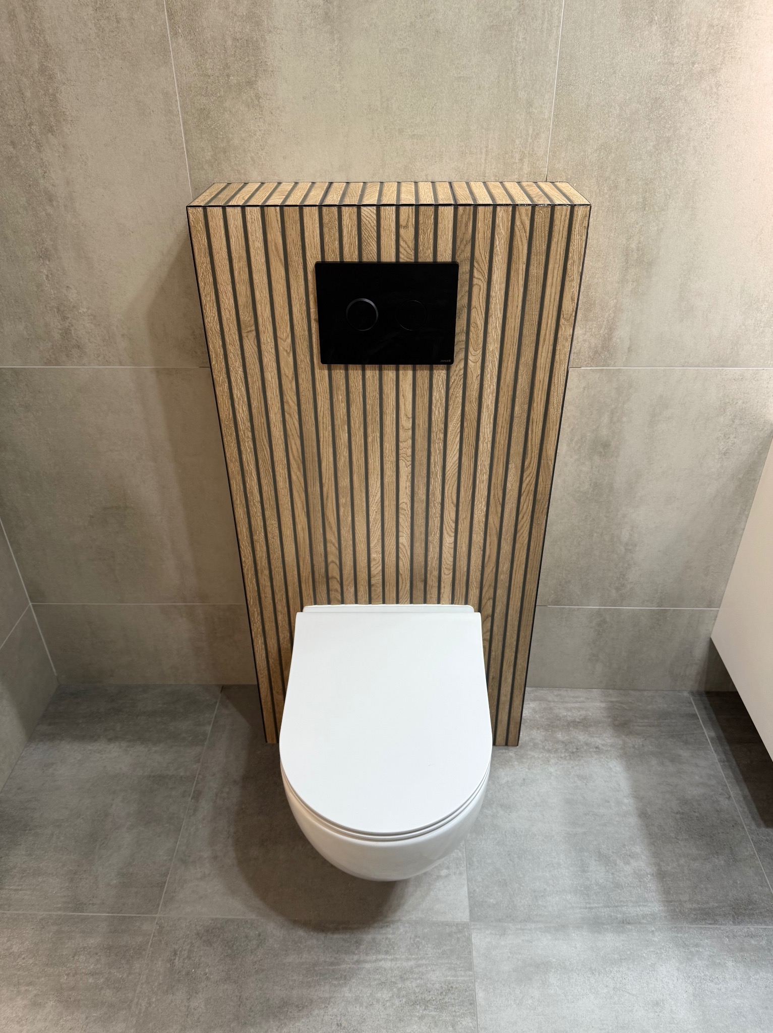 Nowoczesna toaleta podwieszana z drewnianą obudową i czarnym panelem spłukującym, na tle szarych płytek w łazience. Minimalistyczny design.