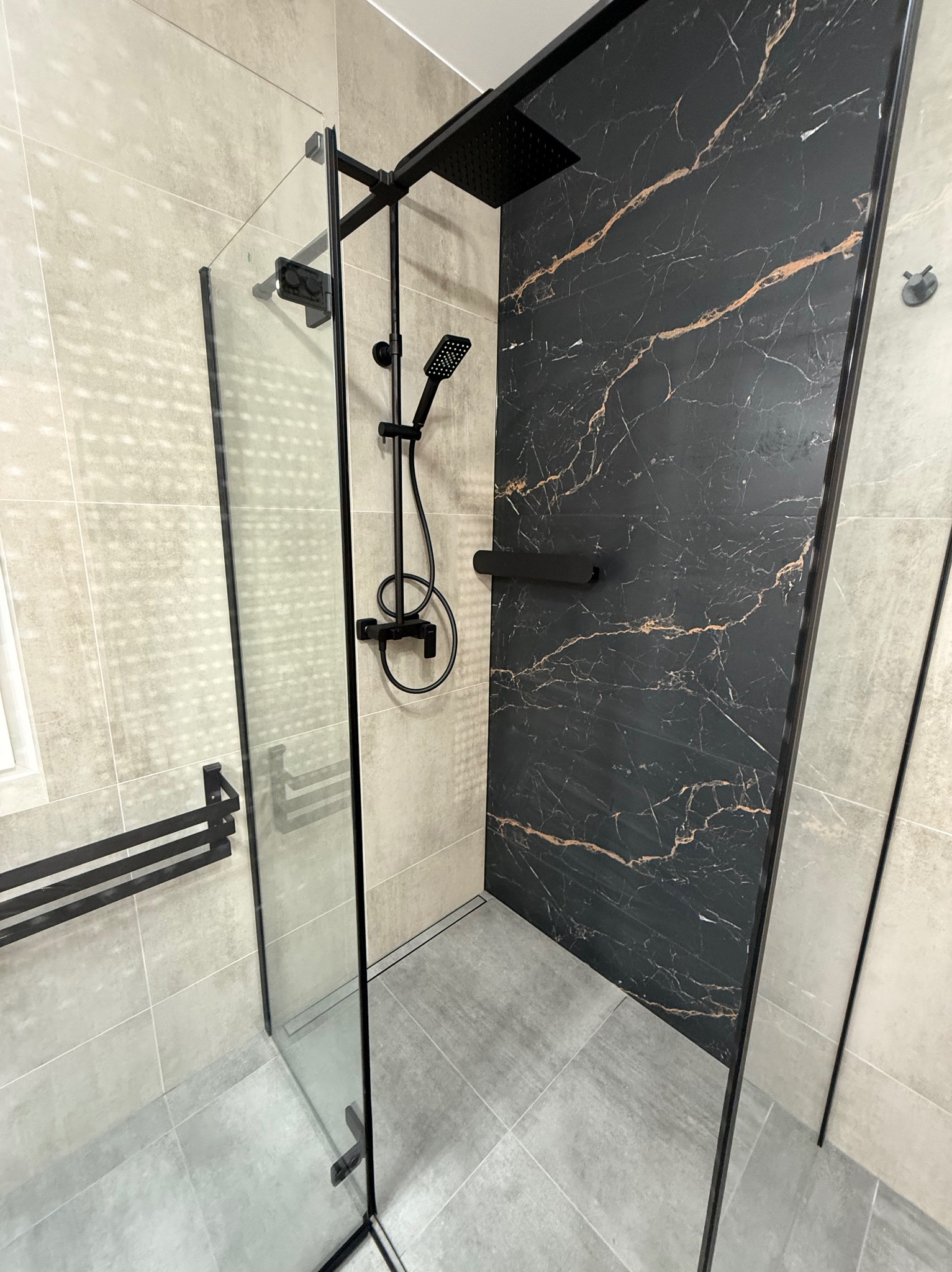 Nowoczesna kabina prysznicowa z czarną armaturą i płytkami imitującymi marmur. Szklane ściany kabiny z czarnymi profilami. Minimalistyczny design łazienki.