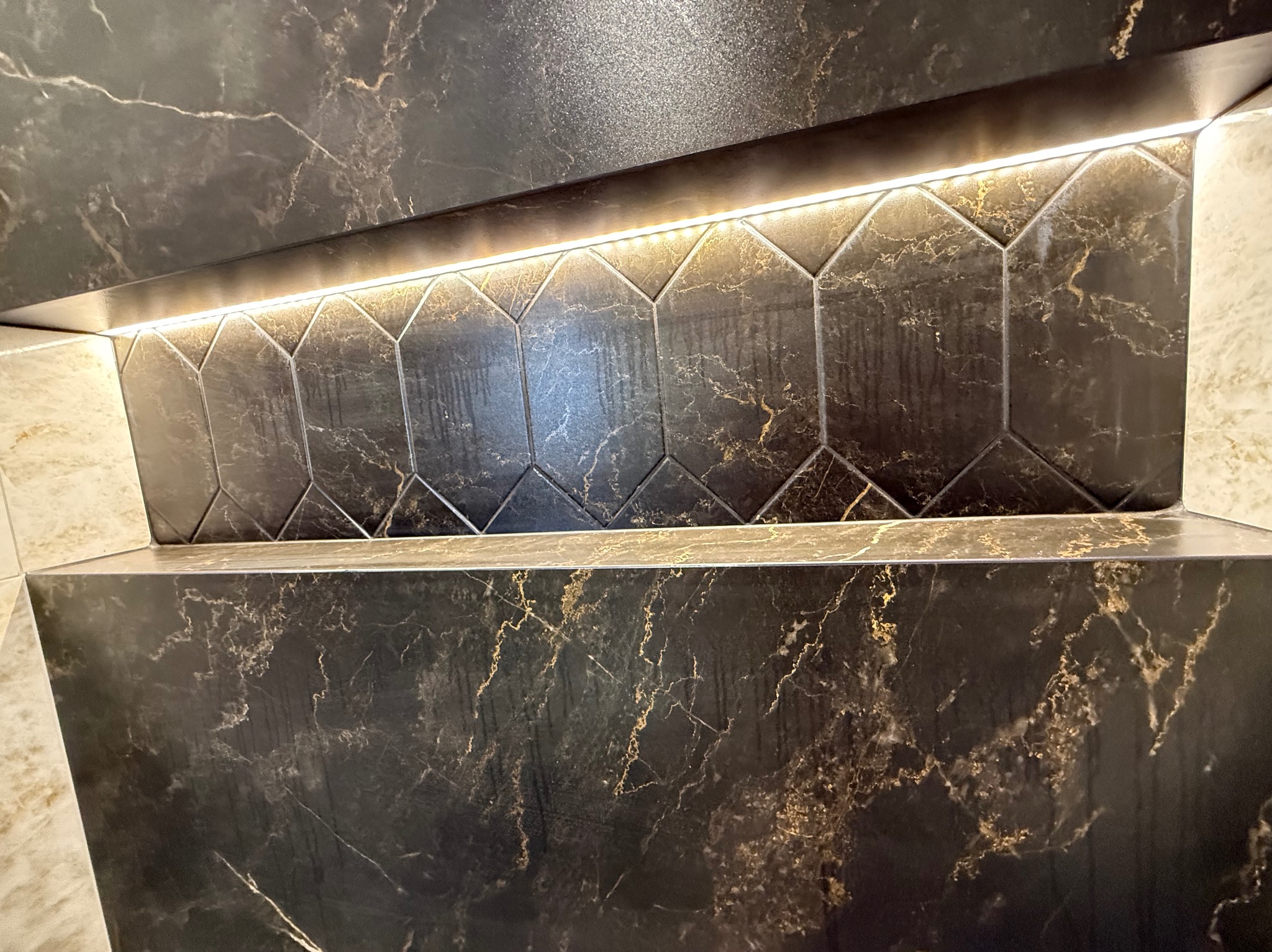 Ciemna, marmurkowa ściana z podświetlaną wnęką wyłożoną heksagonalnymi płytkami. Widoczne złote żyłki w marmurze i precyzyjne fugi.