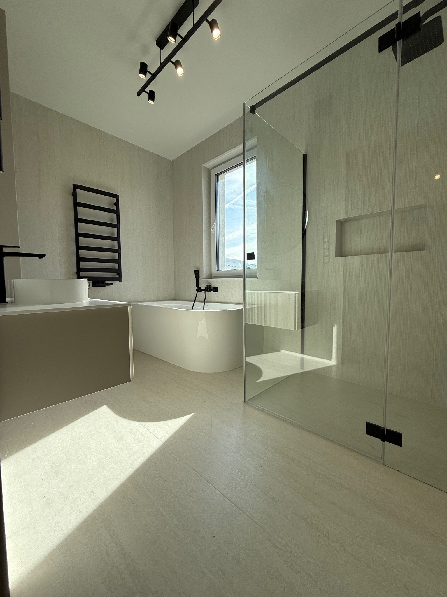 Nowoczesna łazienka z kabiną prysznicową typu walk-in, wolnostojącą wanną i grzejnikiem drabinkowym w czarnym kolorze. Jasne płytki i oświetlenie punktowe.