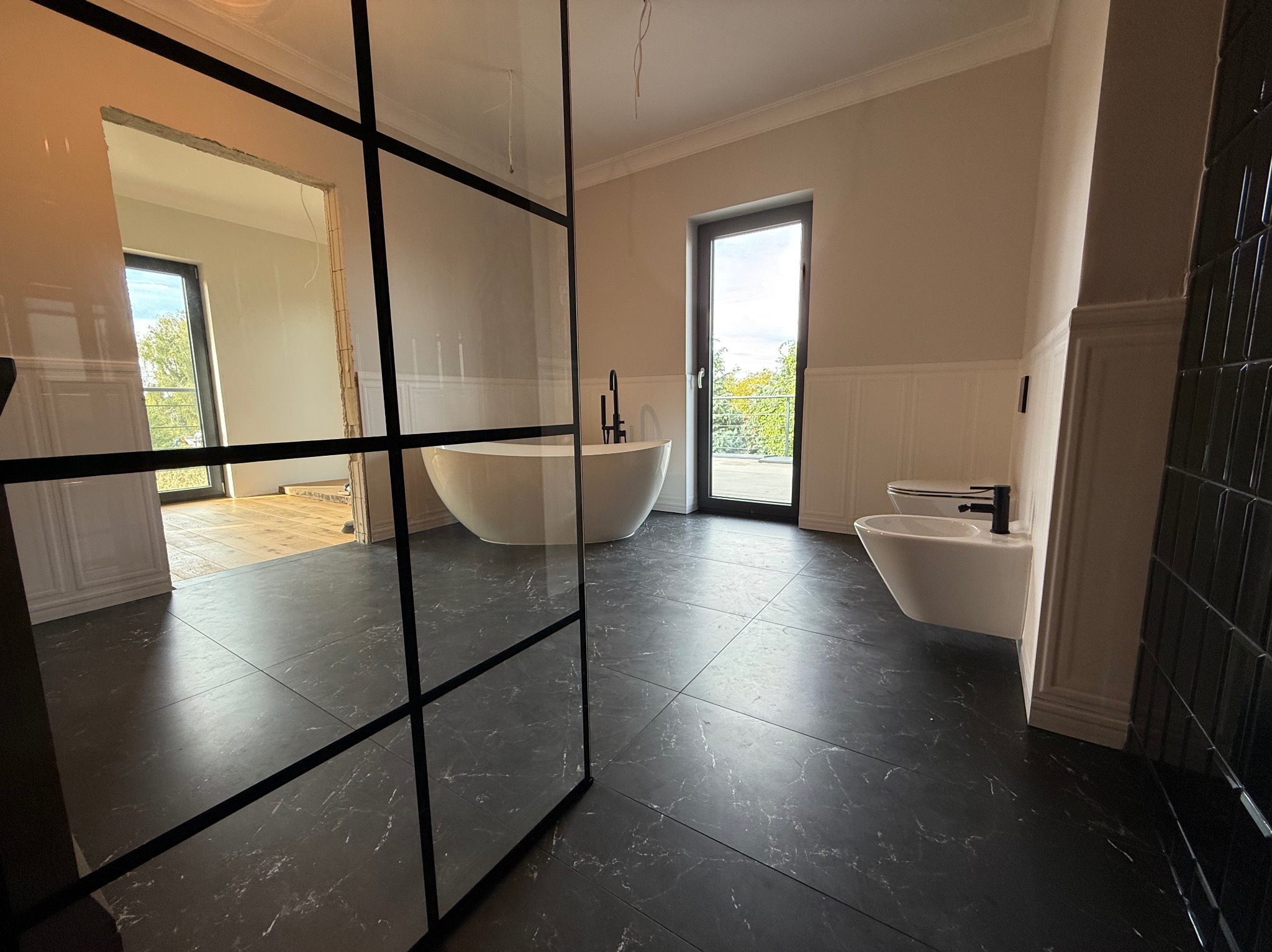 Nowoczesna łazienka z czarną marmurową podłogą, białą wanną wolnostojącą i szklaną ścianką działową z czarnymi profilami. Widok na okno i częściowo wykończone pomieszczenie.