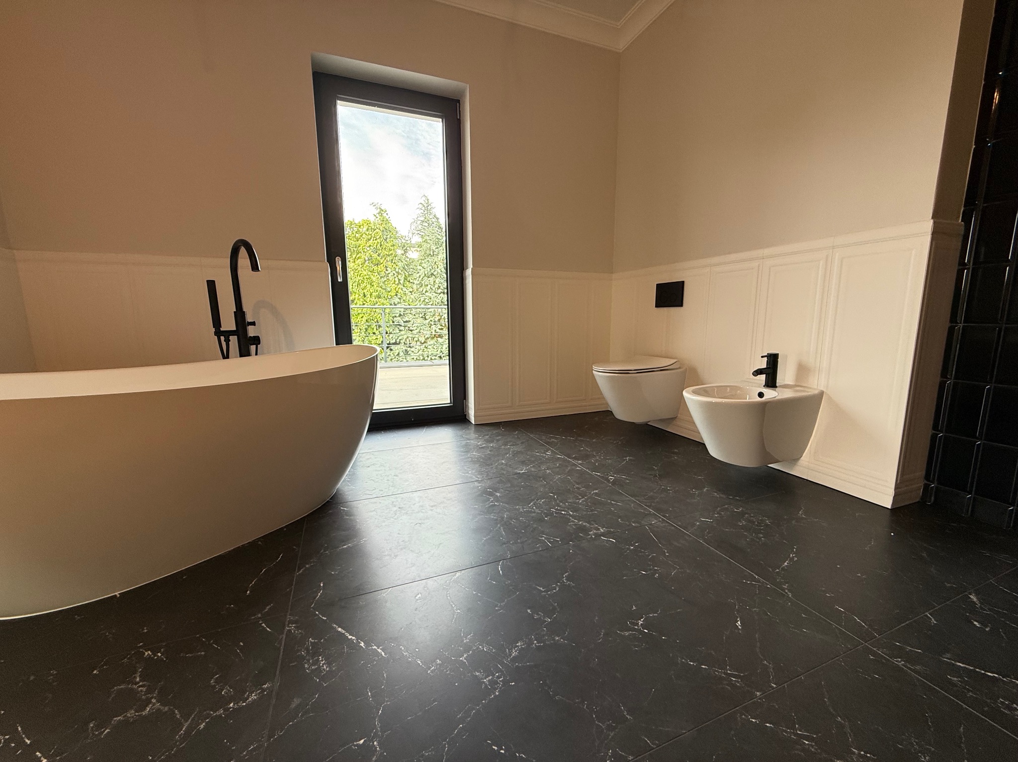 Nowoczesna łazienka z czarną armaturą, marmurową podłogą i wolnostojącą wanną przy oknie z widokiem na zieleń. Minimalistyczny design, eleganckie wykończenie.