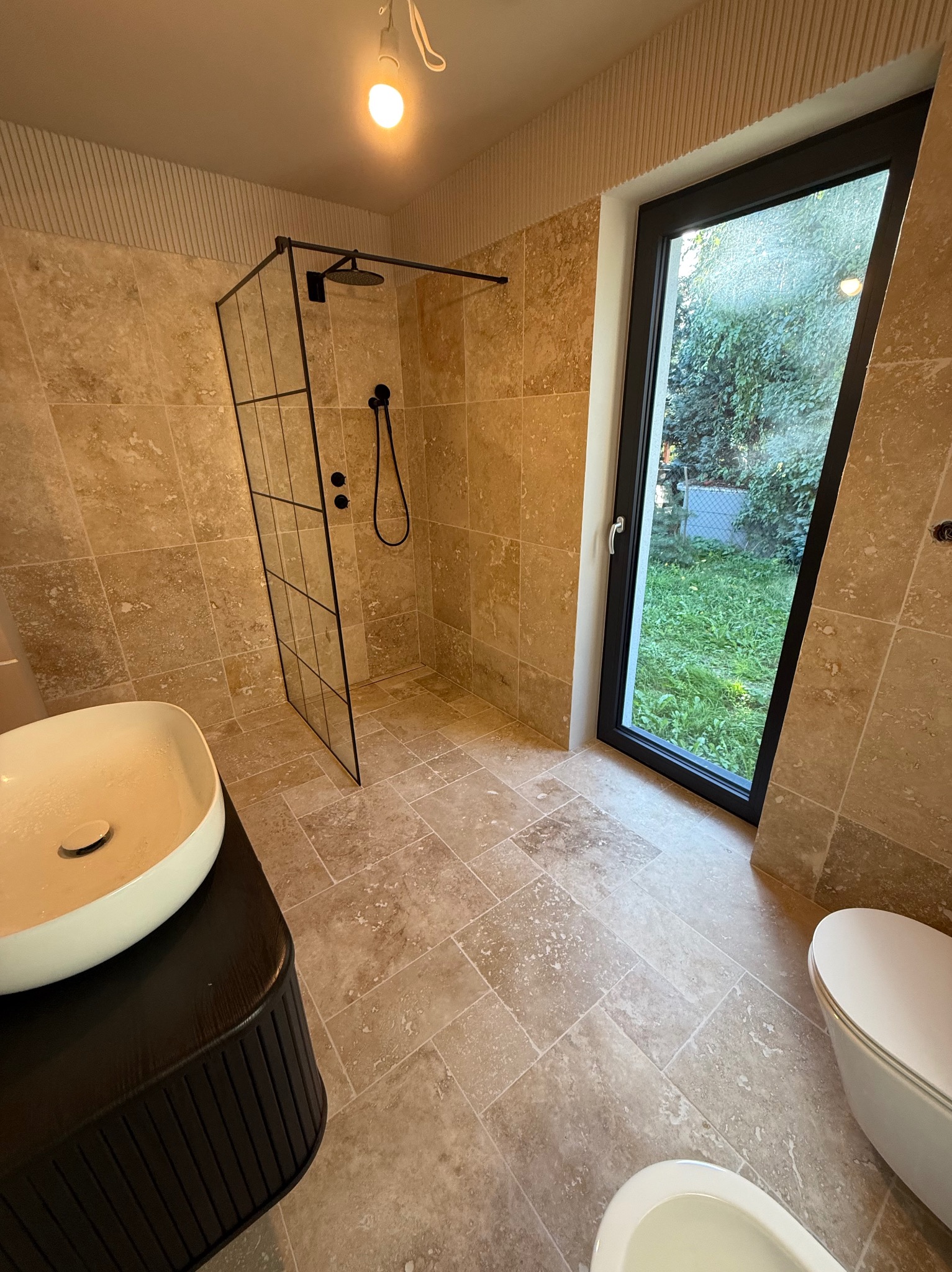 Jasna łazienka z kabiną prysznicową typu walk-in, beżowymi płytkami i czarną stolarką okienną. Umywalka nablatowa na ciemnej szafce. Widok na zieleń za oknem.