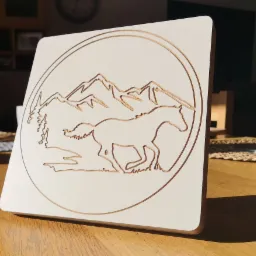 Biała, kwadratowa płyta z wygrawerowanym laserowo motywem konia galopującego na tle gór i drzew, umieszczona na drewnianym stole.