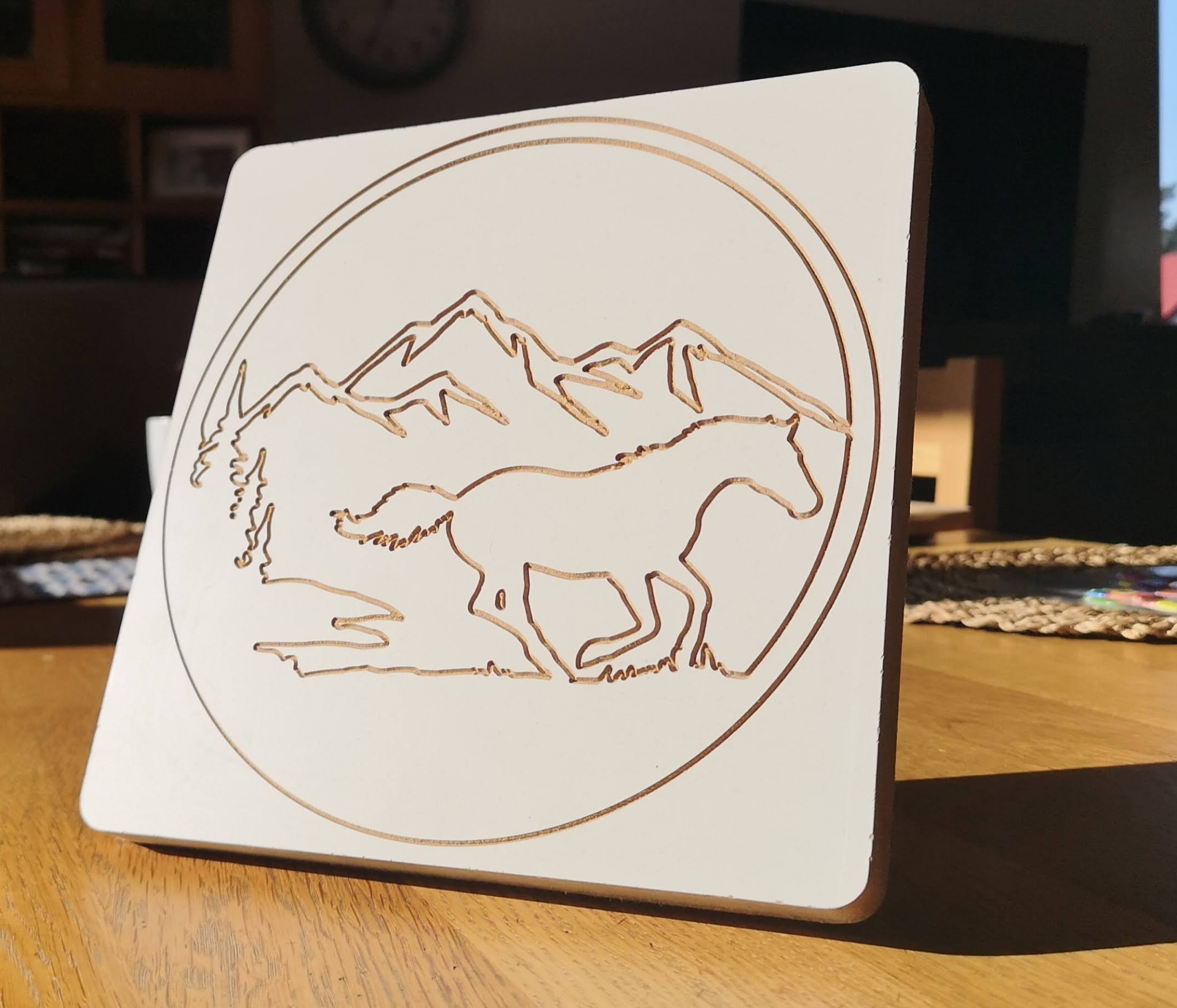 Biała, kwadratowa płyta z wygrawerowanym laserowo motywem konia galopującego na tle gór i drzew, umieszczona na drewnianym stole.