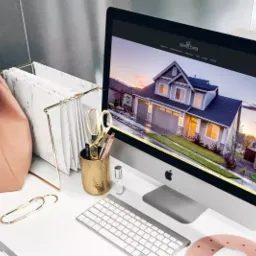 Ekran komputera iMac wyświetlający stronę internetową z wizualizacją domu jednorodzinnego, obok różowy plecak, złoty pojemnik na długopisy i kawa mrożona na białym biurku.