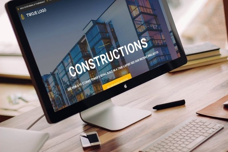 Monitor iMac wyświetlający stronę internetową z dużym napisem 'CONSTRUCTIONS' i wizualizacją nowoczesnego budynku, na biurku leżą klawiatura, smartfon i rysik.