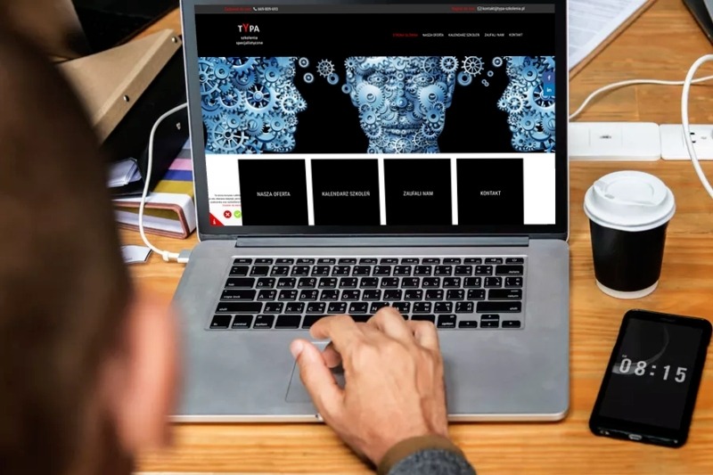 Osoba przegląda na laptopie stronę internetową z motywem ludzkiej głowy zbudowanej z trybików, obok stoi telefon z wyświetlonym czasem 08:15 i kubek z kawą.