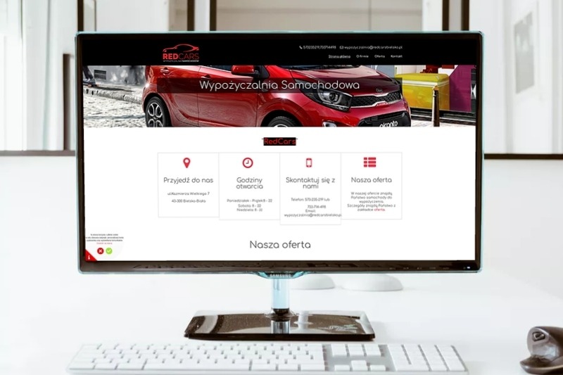 Monitor wyświetla stronę internetową firmy RedCars oferującej wynajem samochodów, widoczny czerwony samochód w banerze, czysty biały pulpit i klawiatura.