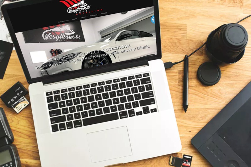 Laptop wyświetlający stronę internetową firmy detailingowej Wasylkowski, obok tablet graficzny, obiektyw, lampa błyskowa i karty pamięci na drewnianym biurku.