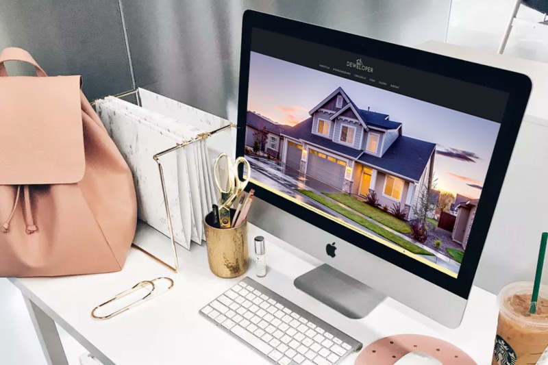 Ekran komputera iMac wyświetlający stronę internetową z wizualizacją domu jednorodzinnego, obok różowy plecak, złoty pojemnik na długopisy i kawa mrożona na białym biurku.