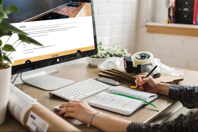 Kobieta notuje w notatniku obok komputera wyświetlającego stronę internetową z projektem kuchni, na biurku akcesoria biurowe i roślina doniczkowa.