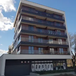 Nowoczesny budynek wielorodzinny z balkonami z brązową elewacją i szklanymi balustradami, z lokalami usługowymi na parterze ozdobionymi sylwetkami kobiecymi, garaż wbudowany w bryłę budynku.