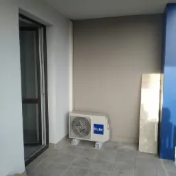 Biała jednostka zewnętrzna klimatyzacji Haier zamontowana na balkonie, widoczne płytki podłogowe i ściana w odcieniach szarości, obok oparta płyta marmurowa.