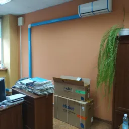 Zamontowana klimatyzacja Haier w biurze, zabezpieczona niebieską taśmą malarską, z niebieskimi rurami odprowadzającymi, obok biurko z stertą dokumentów i kartony z klimatyzatorem na podłodze.