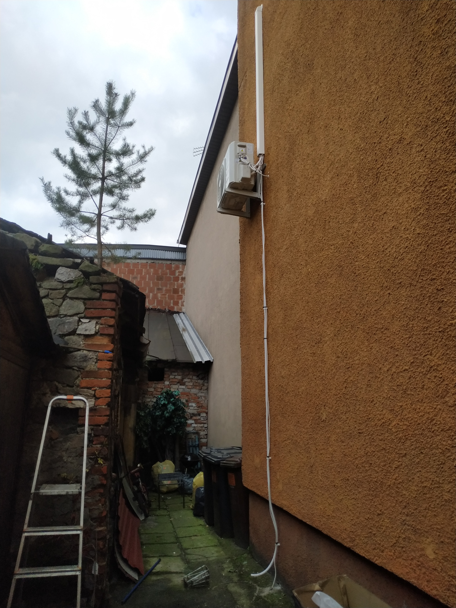 Zewnętrzna jednostka klimatyzacji zamontowana na brązowej ścianie budynku, widoczne kable i antena, w tle drzewo iglaste i fragmenty innych budynków.