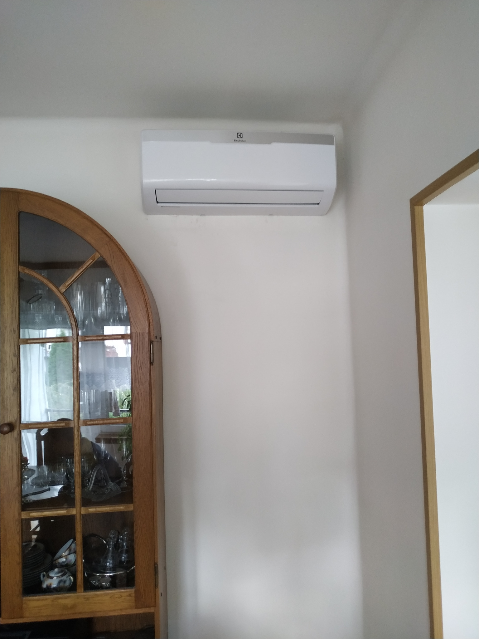 Biała klimatyzacja Electrolux zamontowana na ścianie w pomieszczeniu, widoczna obok drewniana witryna z szklanymi drzwiczkami.