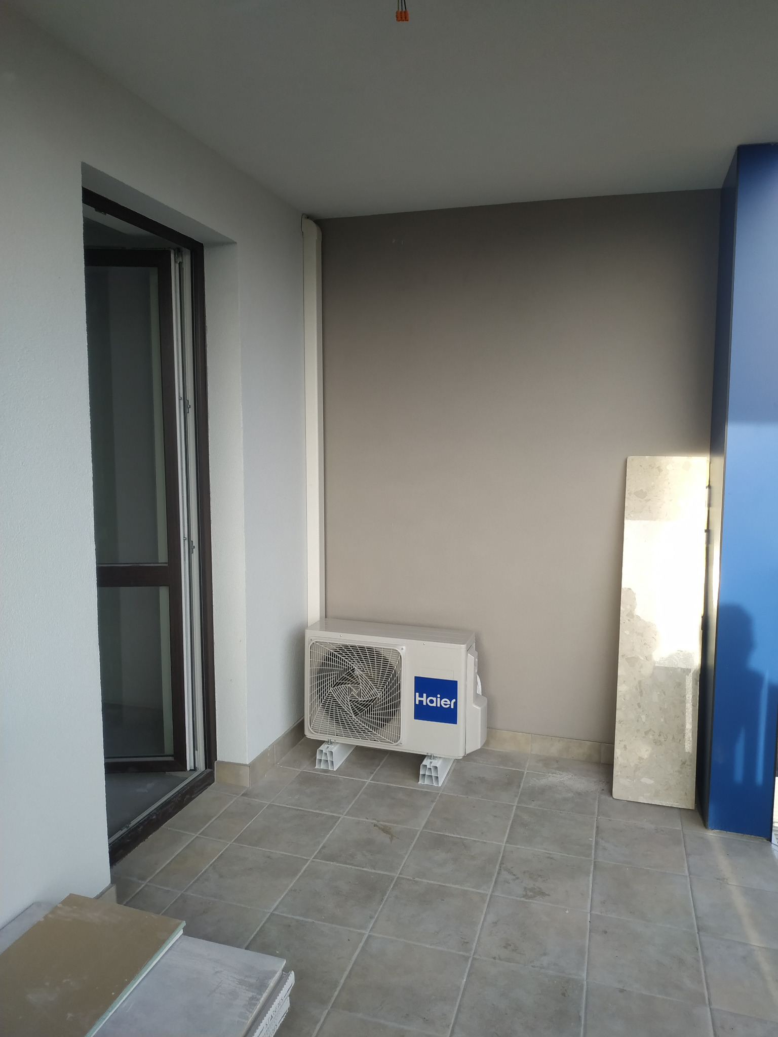 Biała jednostka zewnętrzna klimatyzacji Haier zamontowana na balkonie, widoczne płytki podłogowe i ściana w odcieniach szarości, obok oparta płyta marmurowa.
