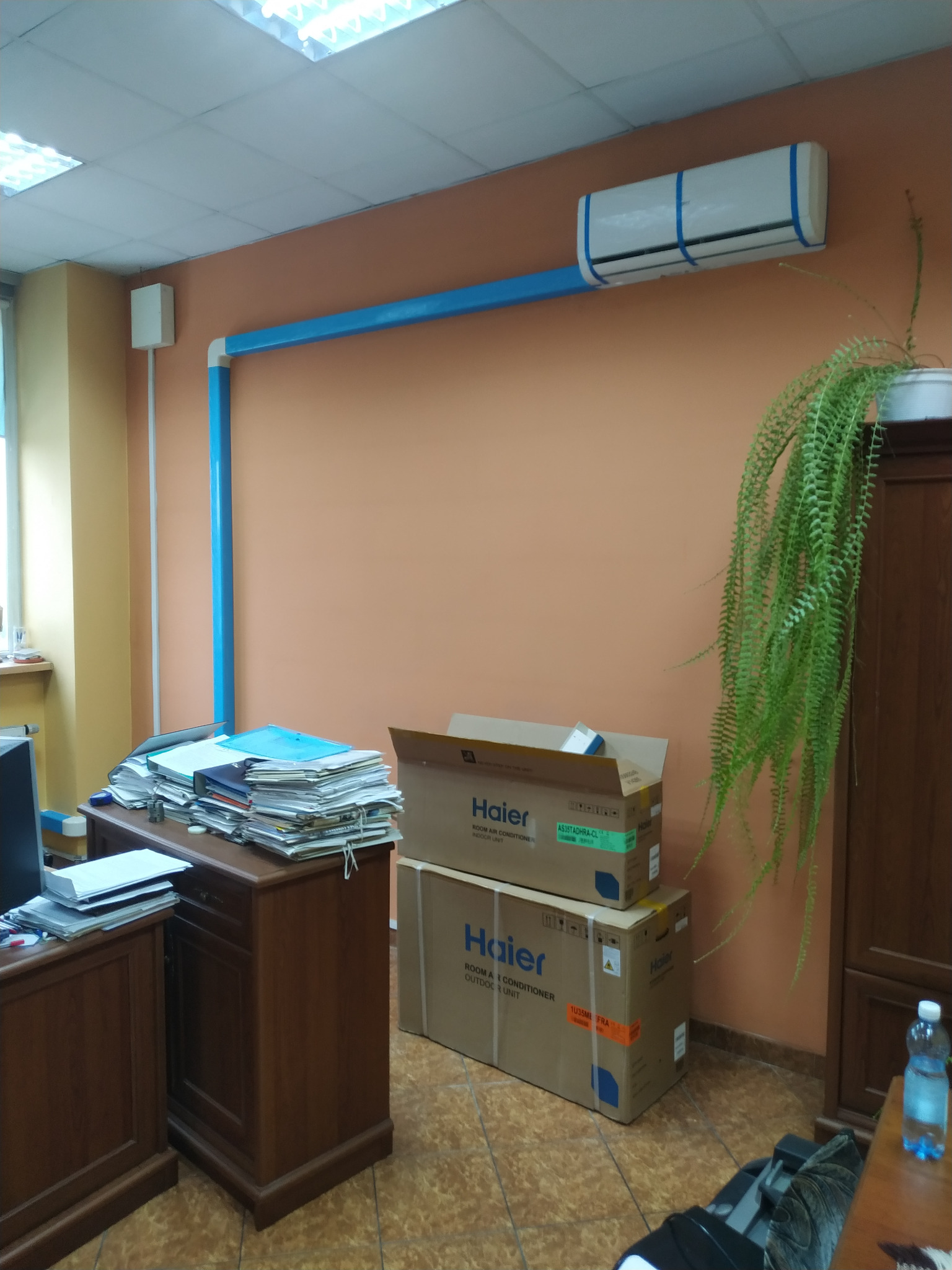 Zamontowana klimatyzacja Haier w biurze, zabezpieczona niebieską taśmą malarską, z niebieskimi rurami odprowadzającymi, obok biurko z stertą dokumentów i kartony z klimatyzatorem na podłodze.