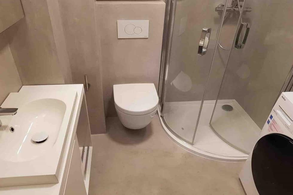 Nowoczesna łazienka z umywalką na szafce, podwieszaną toaletą z przyciskiem spłukującym na ścianie i kabiną prysznicową z zaokrąglonymi drzwiami, wykończona w odcieniach szarości.