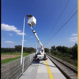 Pracownik w koszu podnośnika na peronie kolejowym, naprawiający oświetlenie uliczne, widok z tyłu na podnośnik i tory kolejowe.