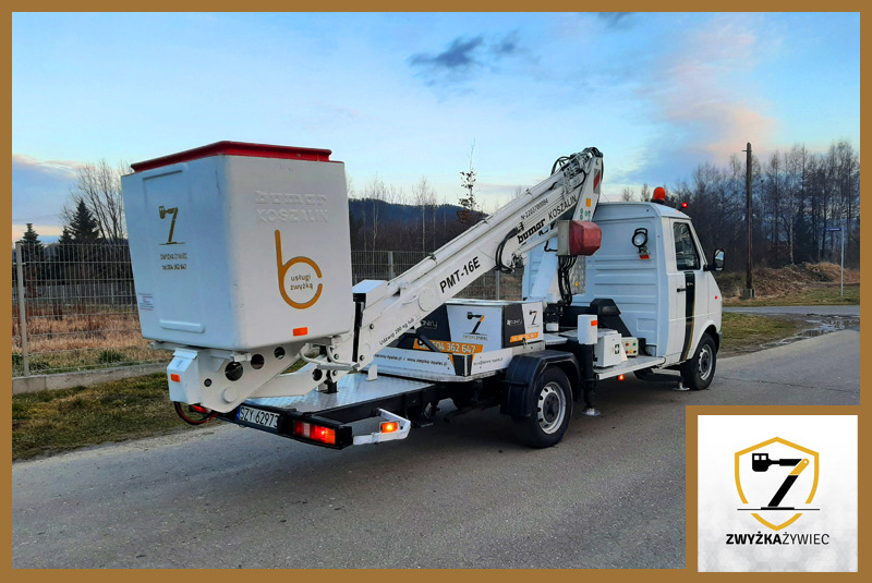 Biały samochód ciężarowy z podnośnikiem koszowym Bumar Koszalin PMT-16E, widok z tyłu, z logo firmy na koszu i nadwoziu, zaparkowany na drodze asfaltowej, w tle drzewa i pagórkowaty krajobraz.