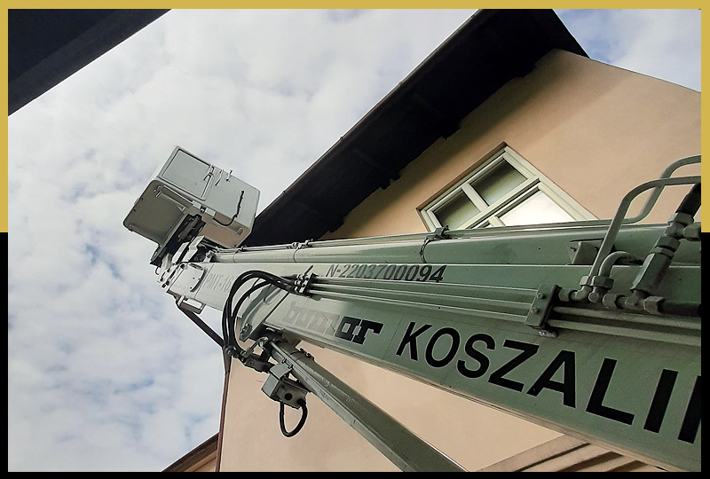 Widok z dołu na wysięgnik z platformą roboczą, z numerem seryjnym N-2203700094, ustawiony przy budynku z beżową elewacją i oknem, na tle pochmurnego nieba.