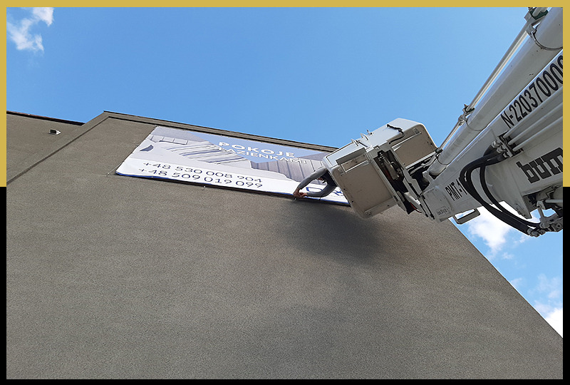 Montaż baneru reklamowego na ścianie budynku z wykorzystaniem podnośnika koszowego, widoczna osoba mocująca baner z bliska.