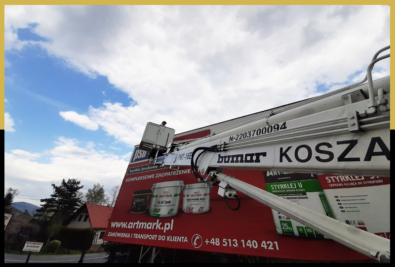 Montaż wielkoformatowej reklamy zewnętrznej z wykorzystaniem podnośnika koszowego na tle błękitnego nieba z chmurami. Widoczny fragment zabudowy miejskiej i drzew.