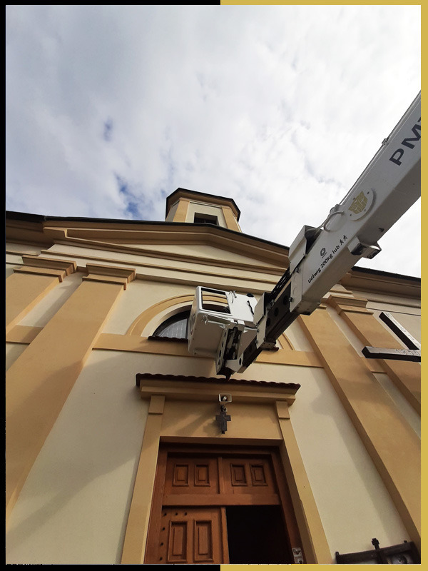 Biały podnośnik z podestem roboczym na tle kościoła z beżową elewacją i drewnianymi drzwiami, widok z dołu na błękitne niebo.