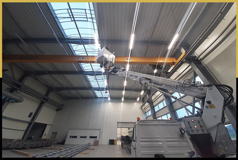 Wnętrze hali produkcyjnej z żółtym suwnicą i białym podnośnikiem koszowym 'Bumar Koszalin' PMT-16E, umieszczającym element pod sufitem.