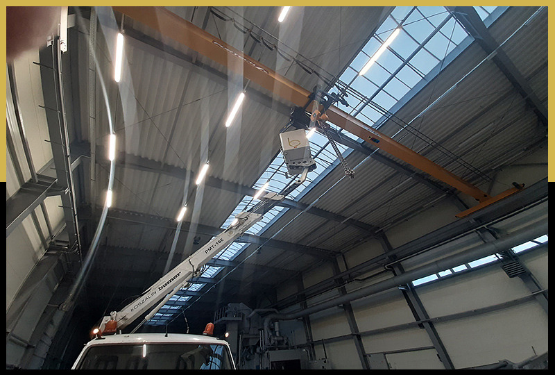 Wnętrze hali przemysłowej z podnośnikiem koszowym teleskopowym podnoszącym element pod sufit, widoczne oświetlenie i konstrukcja dachu.