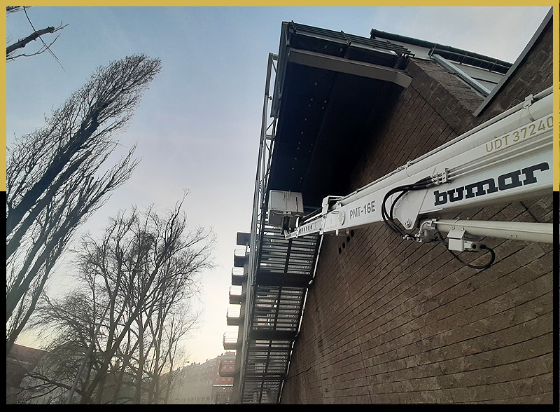 Biały podnośnik marki Bumar z wysięgnikiem PMT-16E przy szarej, ceglanej ścianie budynku, z widocznymi schodami ewakuacyjnymi i drzewami bez liści na tle błękitnego nieba.