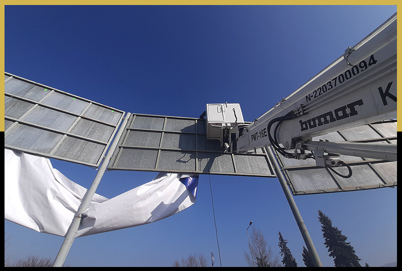 Biały podnośnik koszowy marki Bumar przytwierdzony do tylnej części tablicy reklamowej z resztkami zerwanego baneru na tle błękitnego nieba.