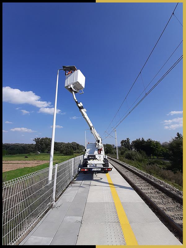 Pracownik w koszu podnośnika na peronie kolejowym, naprawiający oświetlenie uliczne, widok z tyłu na podnośnik i tory kolejowe.