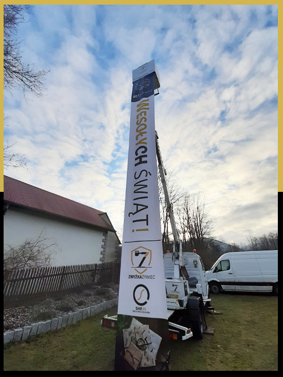 Montaż świątecznej reklamy na wysokim słupie za pomocą pojazdu z wysięgnikiem teleskopowym; widoczny budynek w tle i pochmurne niebo.