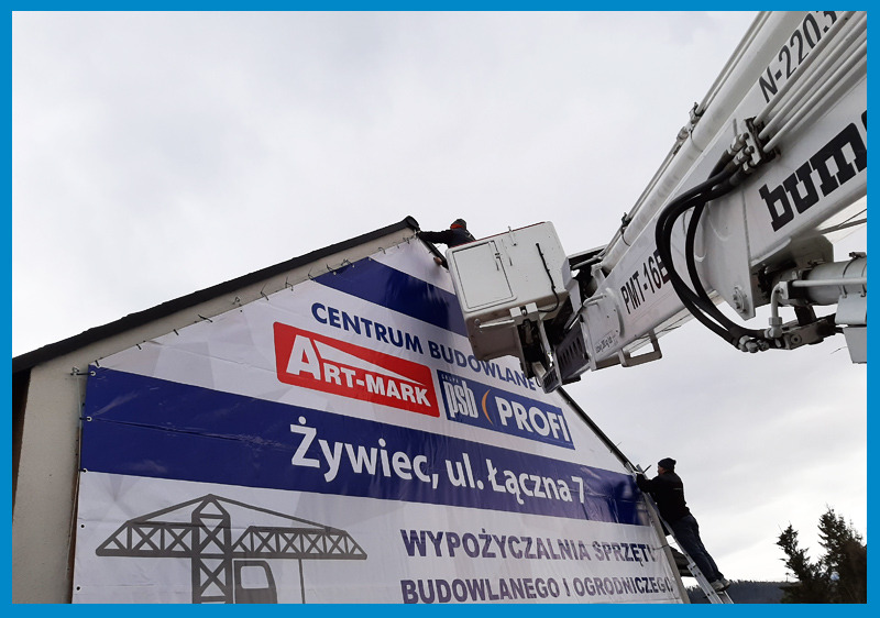 Montaż baneru reklamowego na budynku z wykorzystaniem podnośnika koszowego i drabiny, widoczny fragment elewacji i niebo.