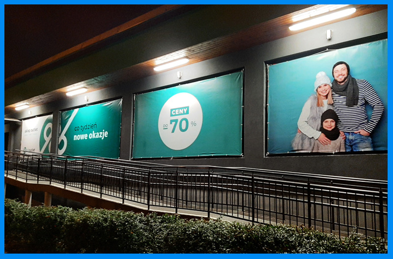 Zewnętrzne banery reklamowe sklepu outletowego z promocjami i zdjęciem rodziny, zamontowane na elewacji budynku przy podjeździe dla wózków.