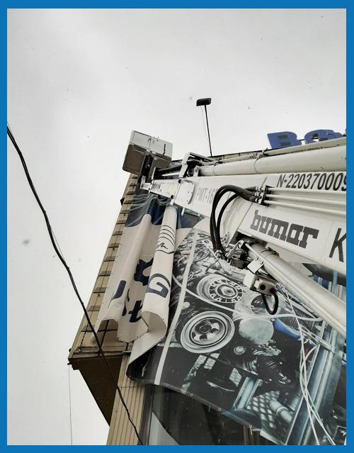 Montaż baneru reklamowego na elewacji budynku z użyciem podnośnika Bumar PMT-16, widok z dołu na pochmurne niebo.