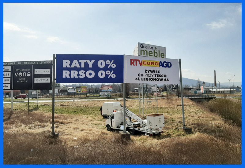 Montaż dwóch dużych billboardów reklamowych z użyciem podnośnika koszowego. Na billboardach widnieją reklamy: 'Raty 0% RRSO 0%' oraz 'Quality meble' i 'RTV Euro AGD Żywiec'. W tle widoczny...