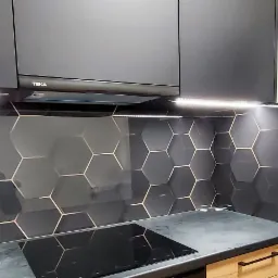 Szklany panel kuchenny w geometryczny wzór heksagonów z miedzianymi obramowaniami, zamontowany pod szafkami z oświetleniem LED i okapem Teka, nad czarną płytą indukcyjną.