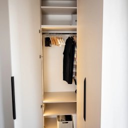 Krzysztof Mędrek - Otwarta szafa wnękowa z oświetleniem LED, widoczne wieszaki z ubraniami w środku i kosz na dole, minimalistyczne czarne uchwyty.