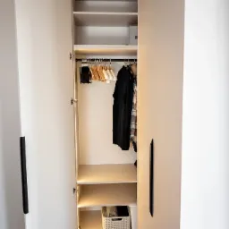 Otwarta szafa wnękowa z oświetleniem LED, widoczne wieszaki z ubraniami w środku i kosz na dole, minimalistyczne czarne uchwyty.