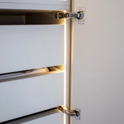 Krzysztof Mędrek - Otwarta szafa z białymi frontami szuflad, oświetleniem LED w pionowych szczelinach i widocznymi zawiasami.